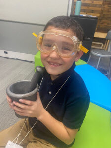 Young MMAEC student smiling at camera with mortar and pestle in the classroom.