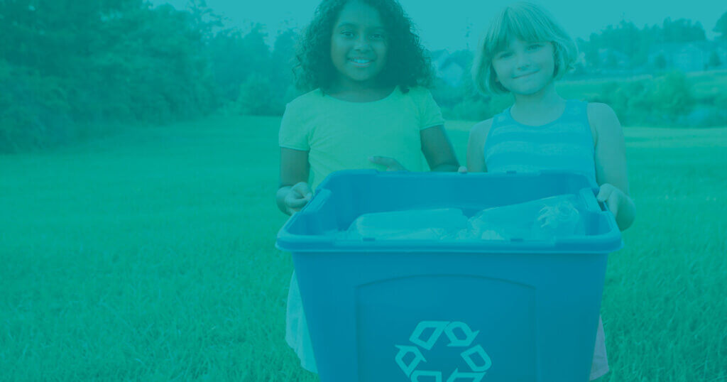 two young students stand in front of a field of grass with a recycling bin